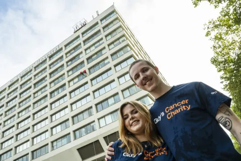 Eddie and Meg smiling at the camera wearing blue Guy's Cancer Charity shirts with St Thomas' building in the background.